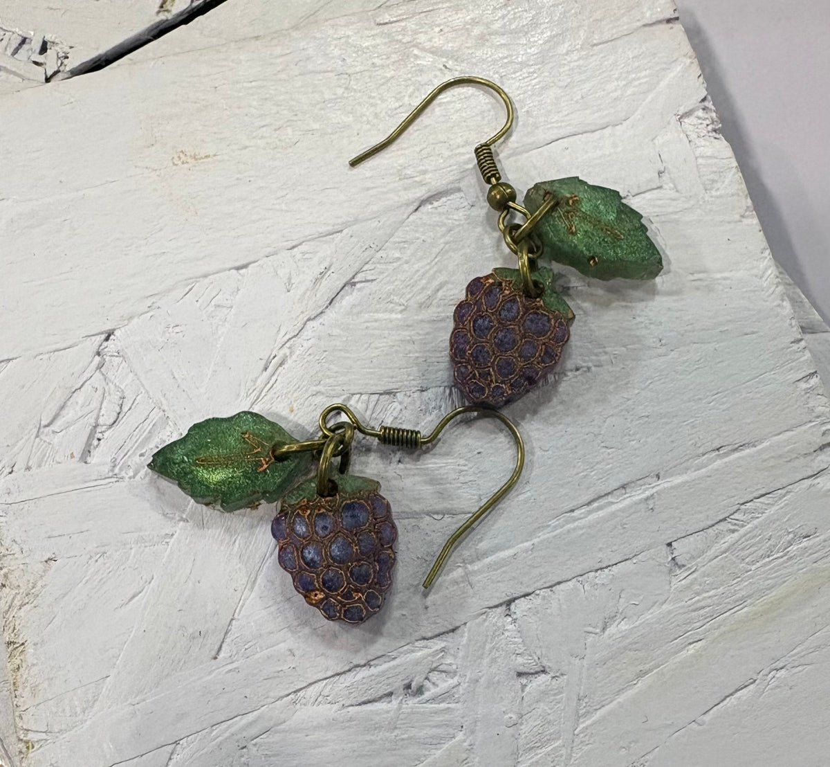 A pair of handmade dangle earrings shaped like purple blackberries with green leaves and gold-tone hooks, displayed on a textured white painted wooden block.