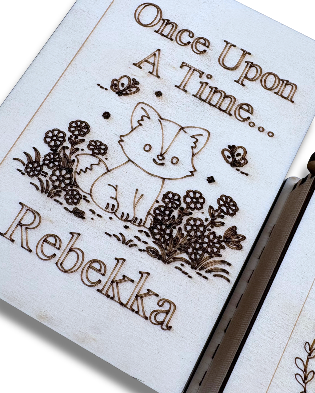 A close-up of a personalized wooden keepsake box or journal page, featuring a laser-engraved illustration of a fox, flowers, butterflies, and the text 'Once Upon A Time.
