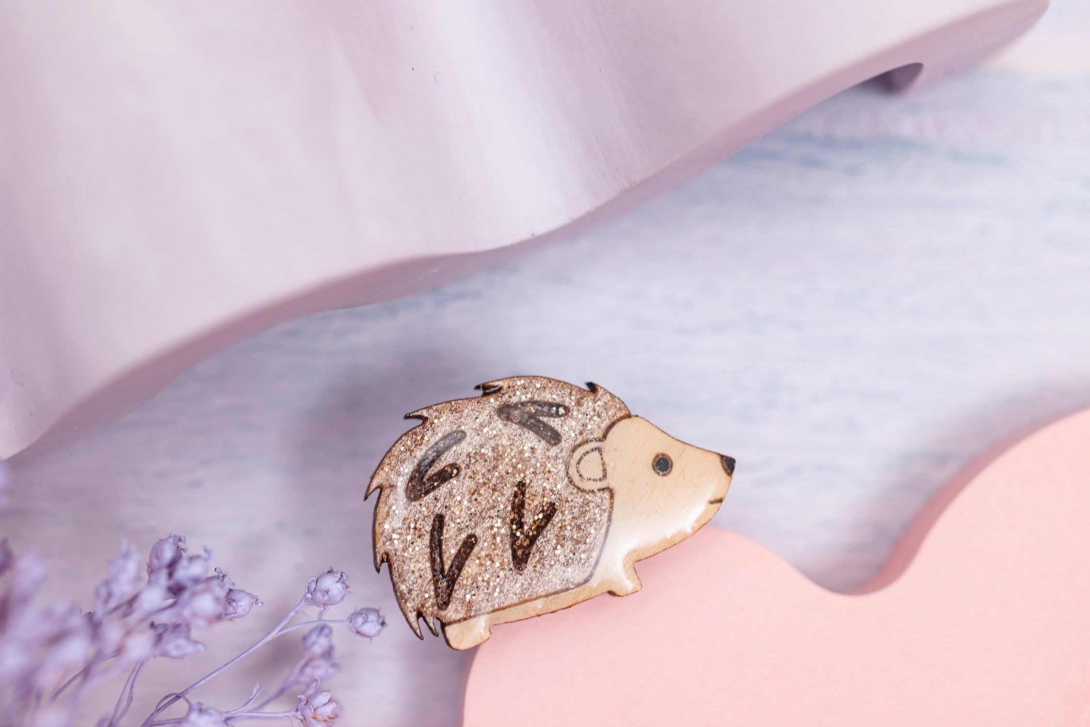 A glittery hedgehog pin badge made of wood, hand-painted and filled with glitter, with a shiny uv resin finish on a pink background.